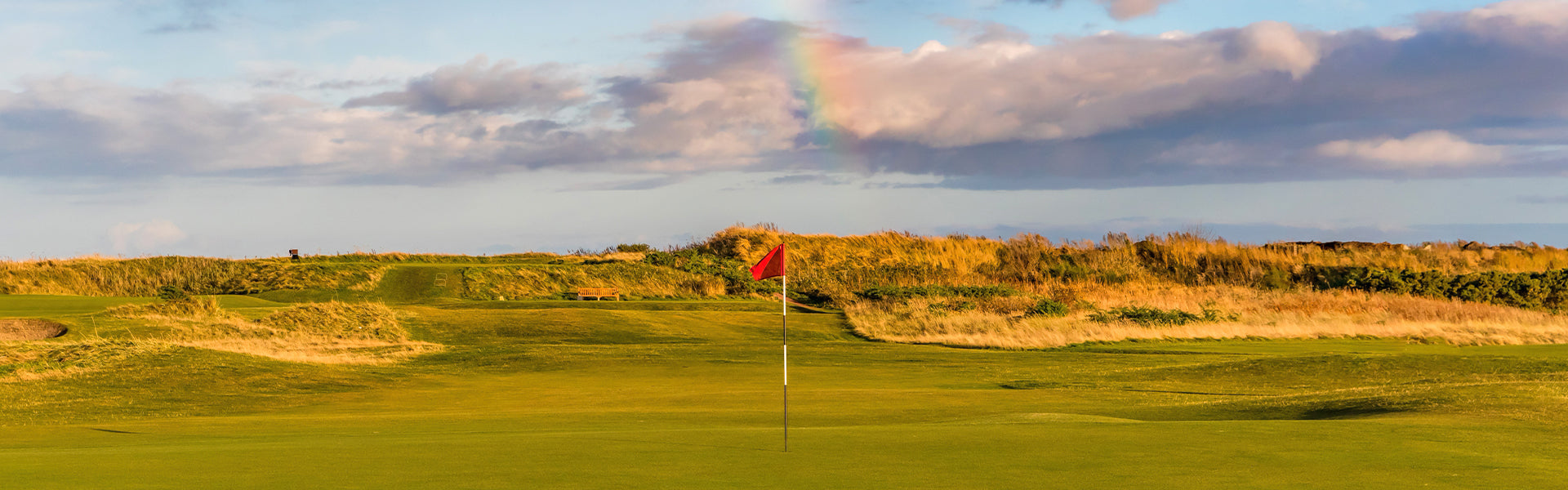 Links-style golf course putting green with a red flag beneath a rainbow and dramatic clouds at sunset.