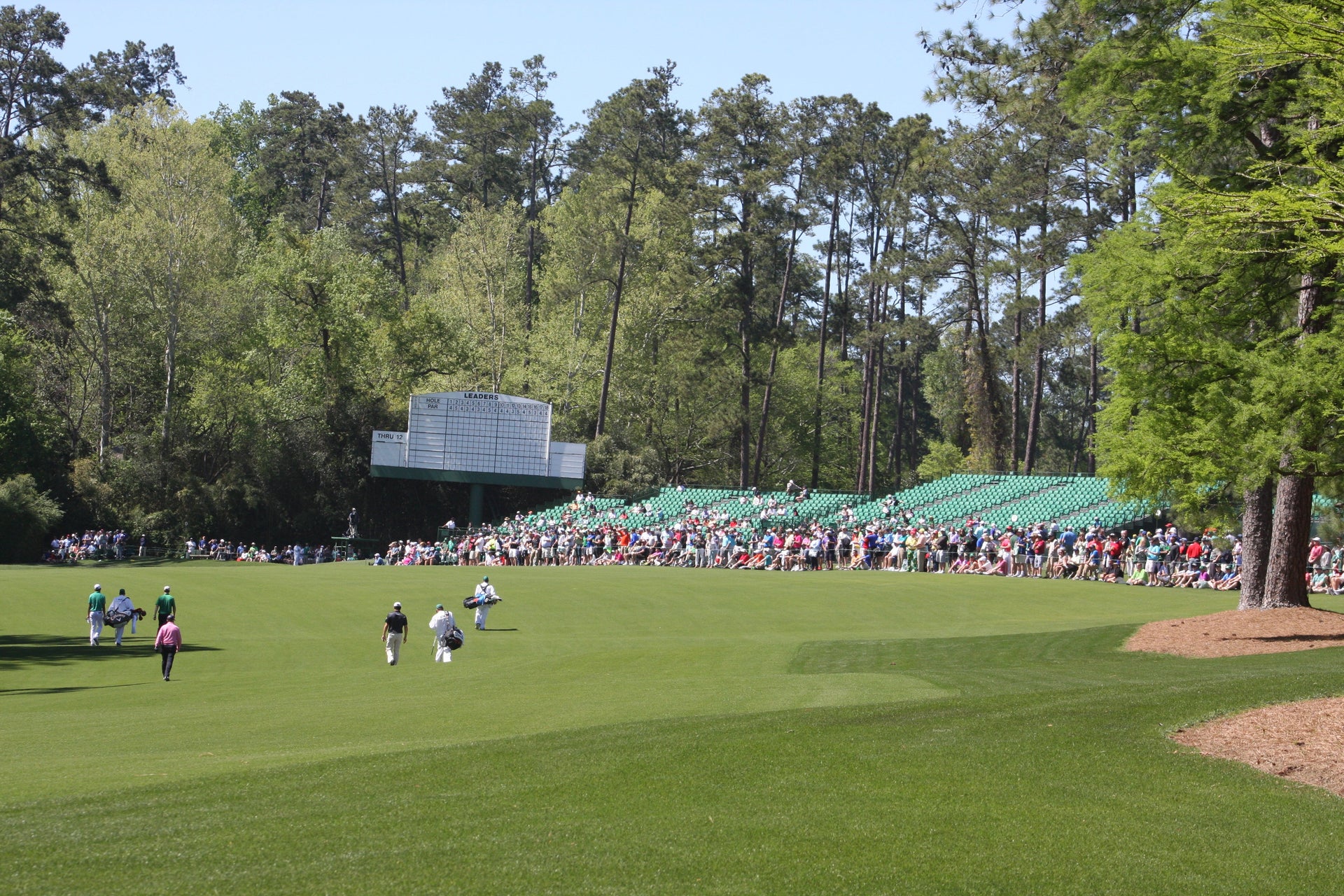 Golfers and caddies walk Augusta National's immaculate fairway past the iconic leaderboard and packed patron grandstands during the Masters Tournament.