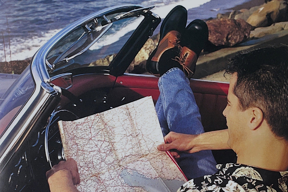 A vintage photo of a man reading a road map with his feet up on the dashboard of a classic convertible parked along the California coast, wearing a floral shirt — a nod to Straight Down's California roots in the 1990s.