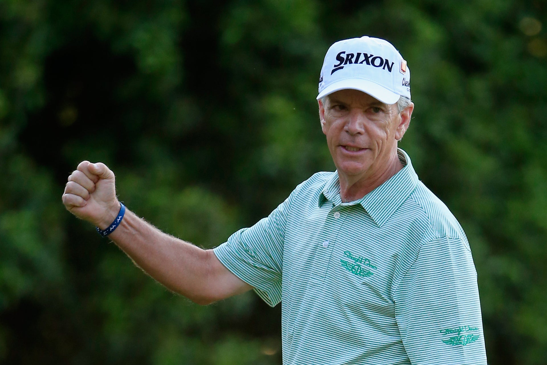 Larry Mize pumps his fist in celebration wearing a Straight Down striped polo and Srixon cap, with a lush green backdrop behind him.