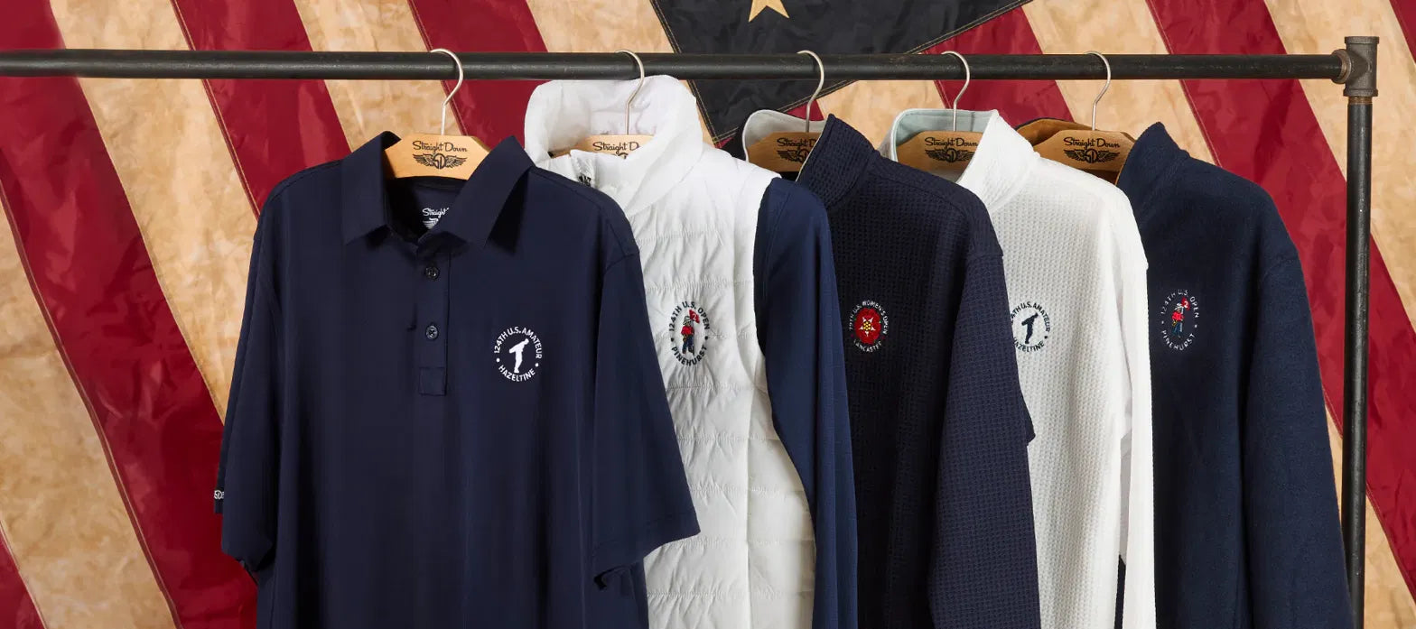 Straight Down apparel featuring embroidered USGA championship logos displayed on a clothing rack with branded wooden hangers against a patriotic flag backdrop.