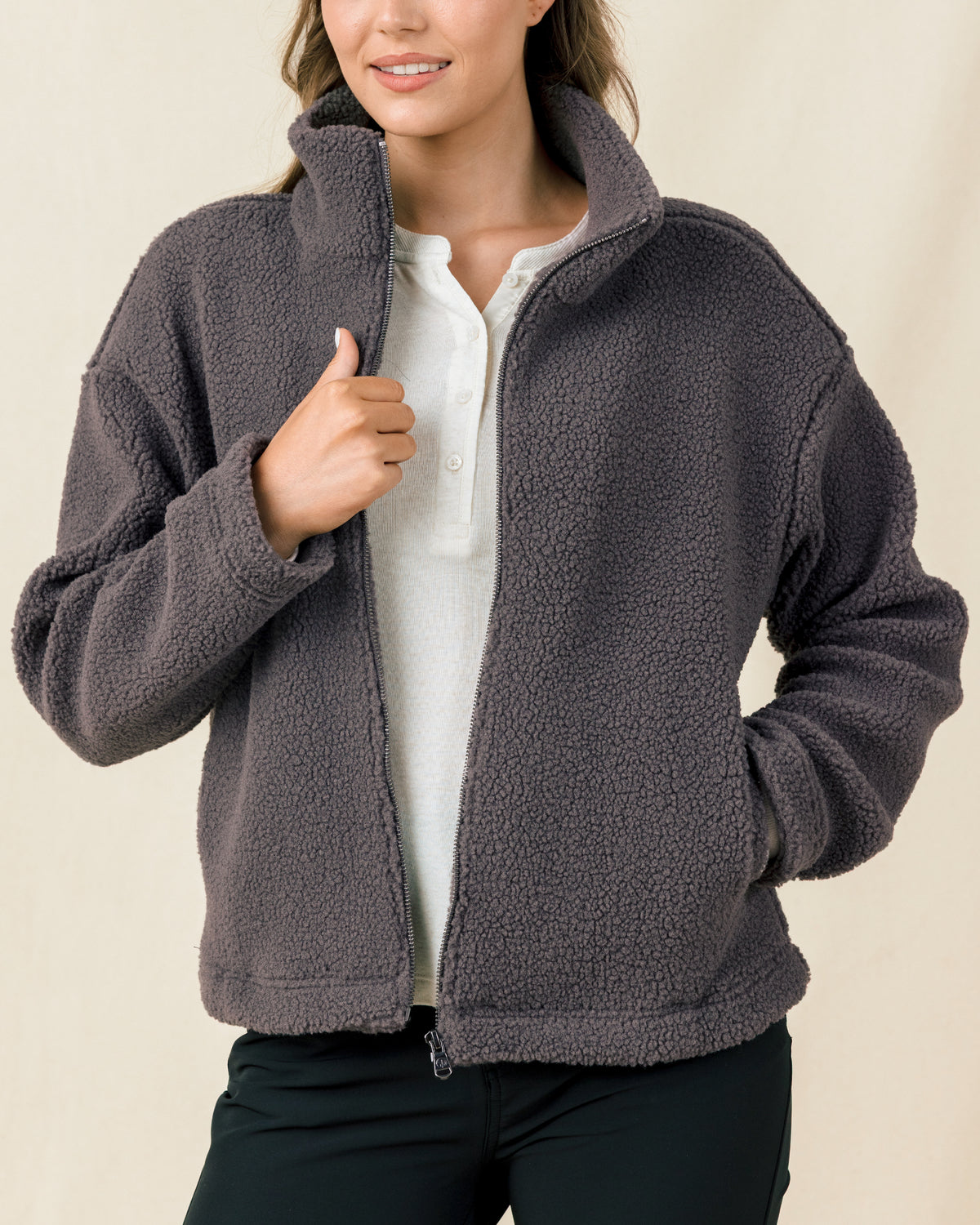A woman modeling a dark gray fleece zip-up jacket over a white shirt and black pants, shown from the nose down against a plain beige studio background.