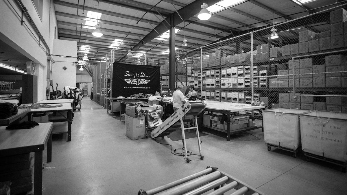Black and white warehouse interior with tall storage racks full of boxes, chain-link fencing, and a busy packing station beneath a banner reading Straight Down