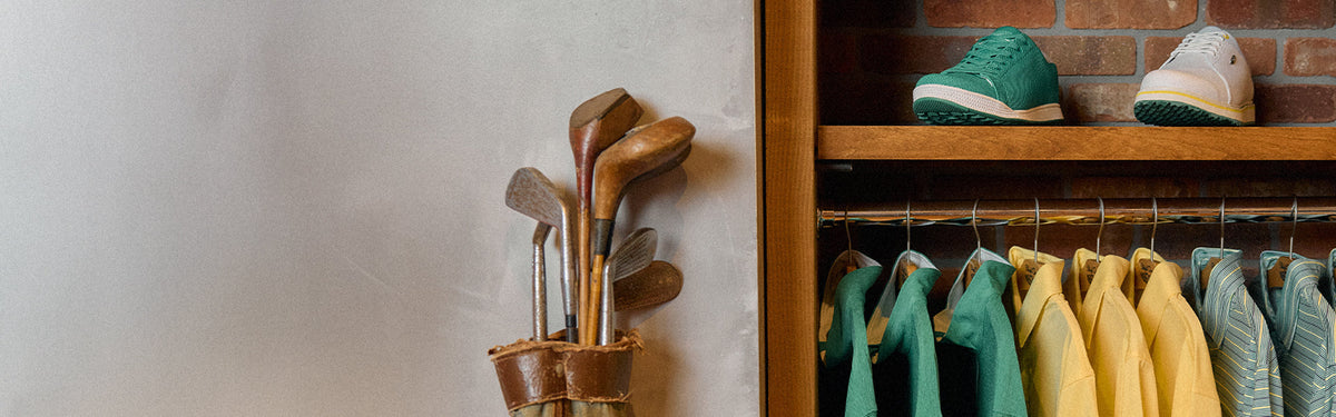 Vintage hickory golf clubs in a leather bag beside a wooden wardrobe displaying green and yellow Straight Down polos and golf shoes, evoking classic Masters Tournament style.