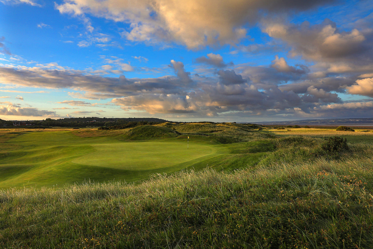  Links golf course green with a yellow flag surrounded by native rough and dunes, with a cloudy sky and coastal landscape stretching to the horizon.