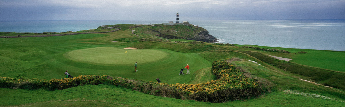 Golfers on a clifftop links green at an iconic Irish coastal course, with a lighthouse and the Atlantic Ocean stretching to the horizon beyond.