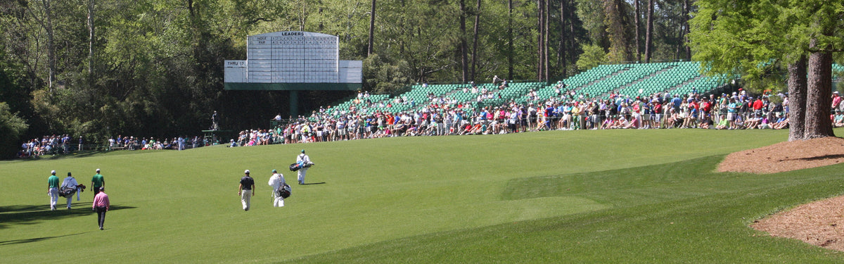 Crowd of spectators lines a sunny golf course as players and caddies walk toward the green with a large scoreboard in the background.