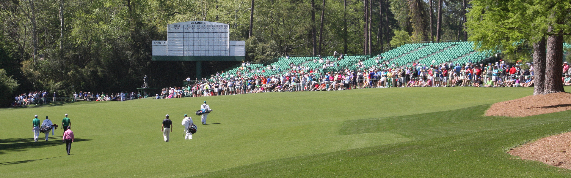 Crowd of spectators lines a sunny golf course as players and caddies walk toward the green with a large scoreboard in the background.