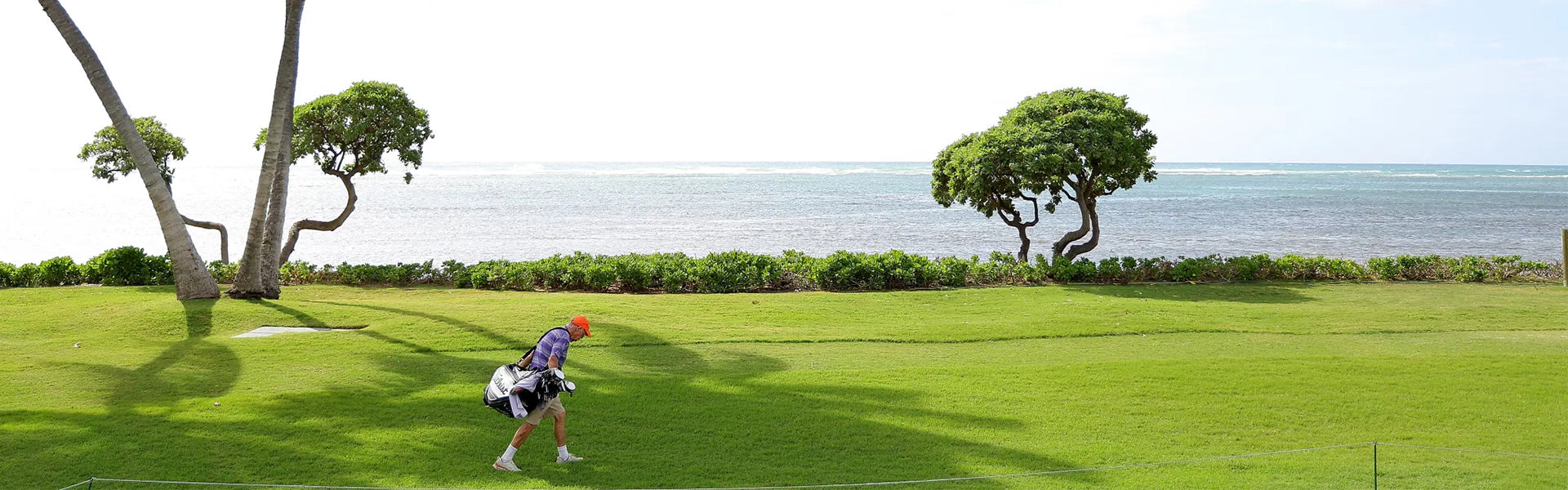 A golfer carrying a bag walks across a lush oceanside fairway with tropical trees and ocean views in the background.
