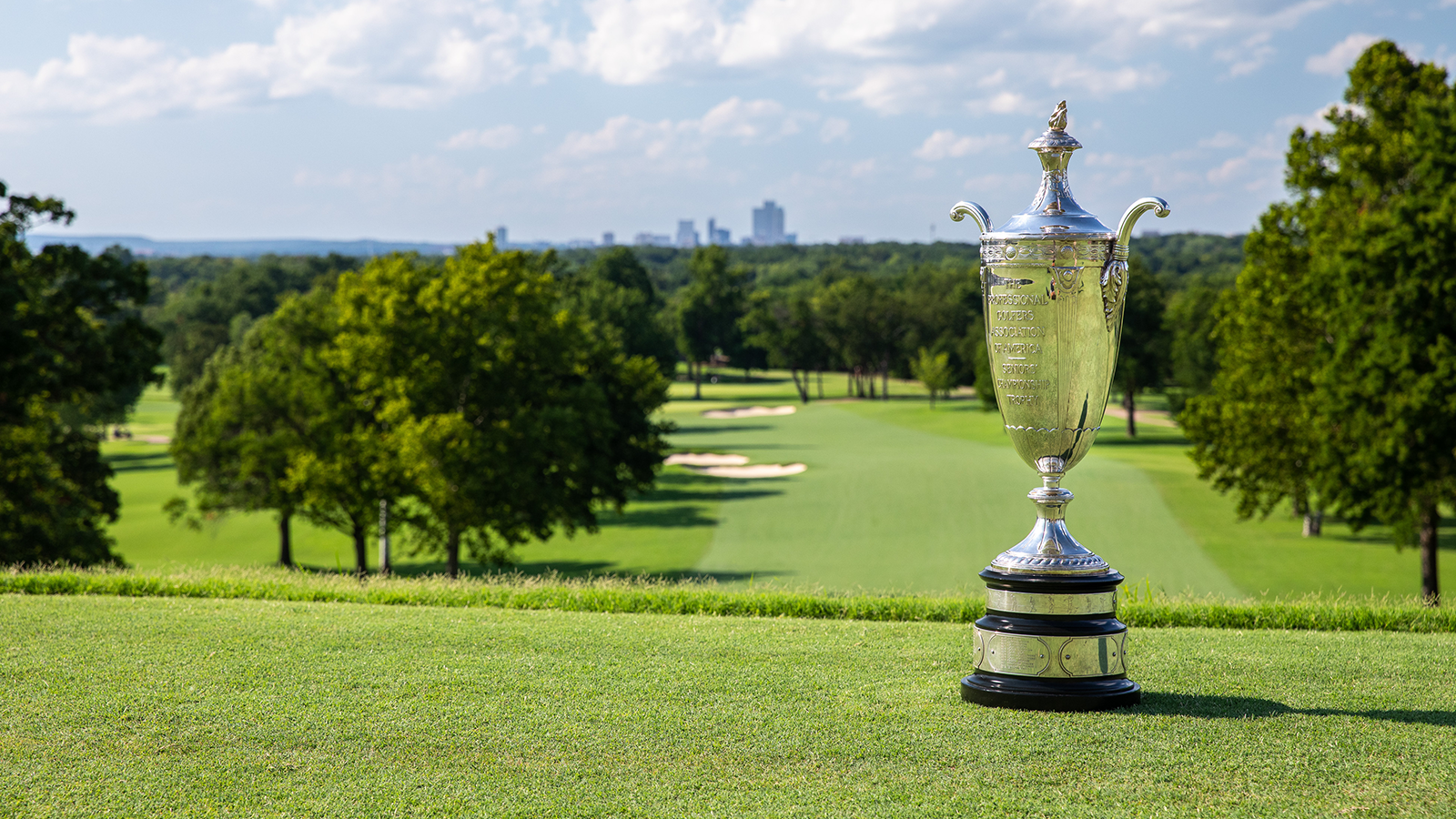 The Wanamaker Trophy sits on the fairway at Colonial Country Club with the Fort Worth skyline visible through the trees in the background.