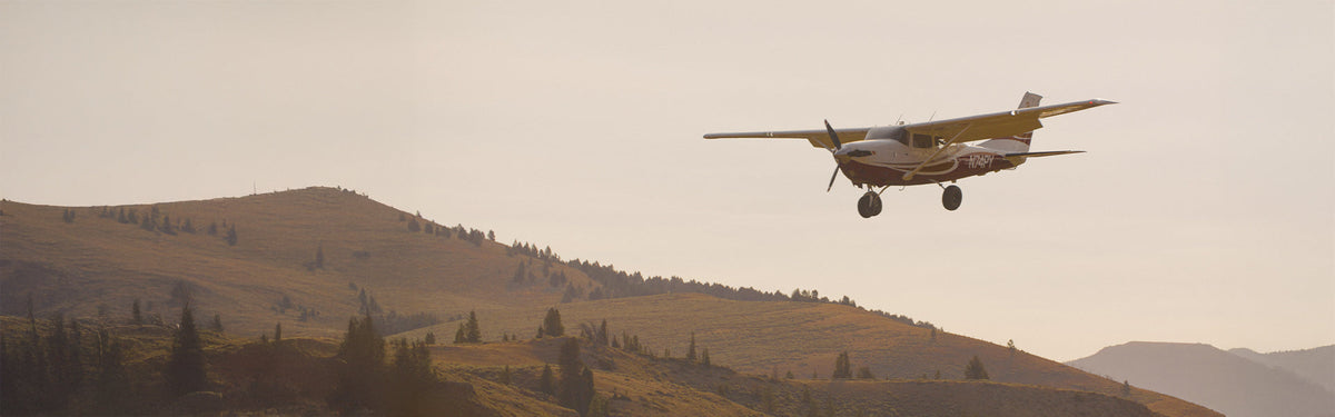 A small propeller plane flies over golden mountain foothills at dusk, evoking the spirit of adventure and travel that inspires Straight Down's golf lifestyle.