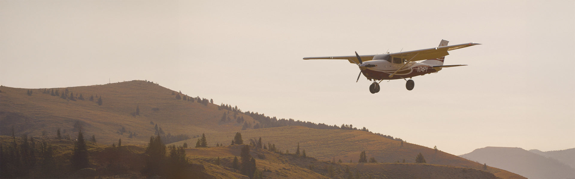 A small propeller plane flies over golden mountain foothills at dusk, evoking the spirit of adventure and travel that inspires Straight Down's golf lifestyle.