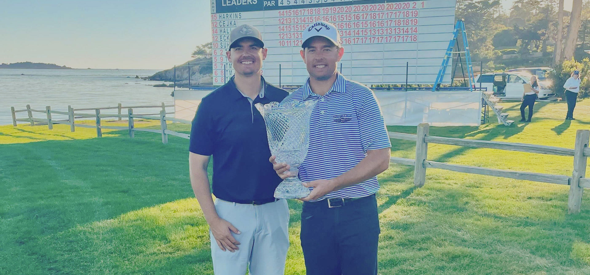 Brandon Harkins holds the Pebble Beach Invitational crystal trophy alongside a companion in front of the tournament leaderboard, wearing a Straight Down striped polo with the Monterey Bay coastline in the background.