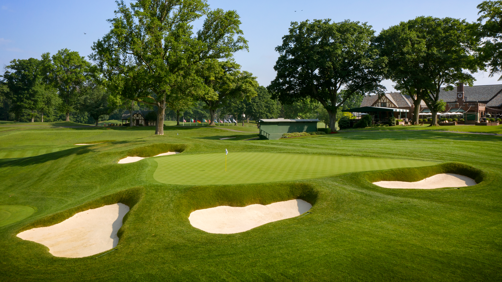 A pristine green surrounded by sculpted sand bunkers at Oak Hill Country Club, with the Tudor-style clubhouse visible through the mature trees in the background.