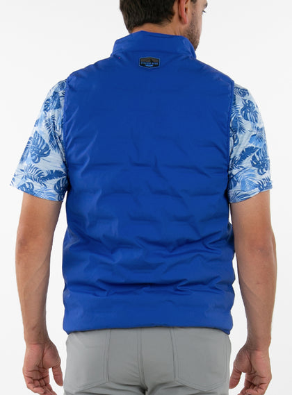 Men's Peak Vest in Azurite, on-model back view featuring a royal blue quilted vest with a branded logo badge at the nape worn over a blue tropical print polo with gray pants