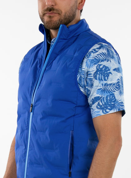 Men's Peak Vest in Azurite, on-model close-up front view featuring a royal blue quilted vest highlighting the mock neck collar, light blue full zip closure, and side zip pocket detail worn over a blue tropical print polo