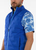 Men's Peak Vest in Azurite, on-model close-up front view featuring a royal blue quilted vest highlighting the mock neck collar, light blue full zip closure, and side zip pocket detail worn over a blue tropical print polo