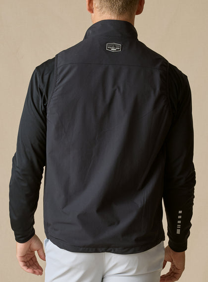 Men's Denver Vest in Black, back model view featuring a black shell with woven logo patch at the upper back, layered over a black long sleeve pullover with light gray pants