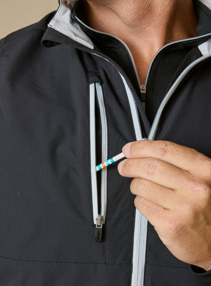 Men's Denver Vest in Black, detail close-up front view featuring a black shell with light gray contrast zippered chest pocket, demonstrating the functional tee storage detail with a golf tee being retrieved