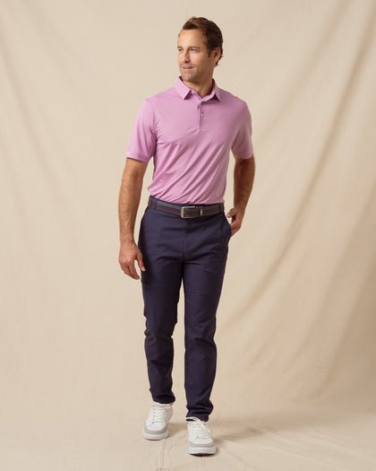 Man wearing a haze pink/purple short-sleeve polo shirt paired with navy pants, a brown belt, and white sneakers against a neutral backdrop
