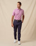Man wearing a haze pink/purple short-sleeve polo shirt paired with navy pants, a brown belt, and white sneakers against a neutral backdrop