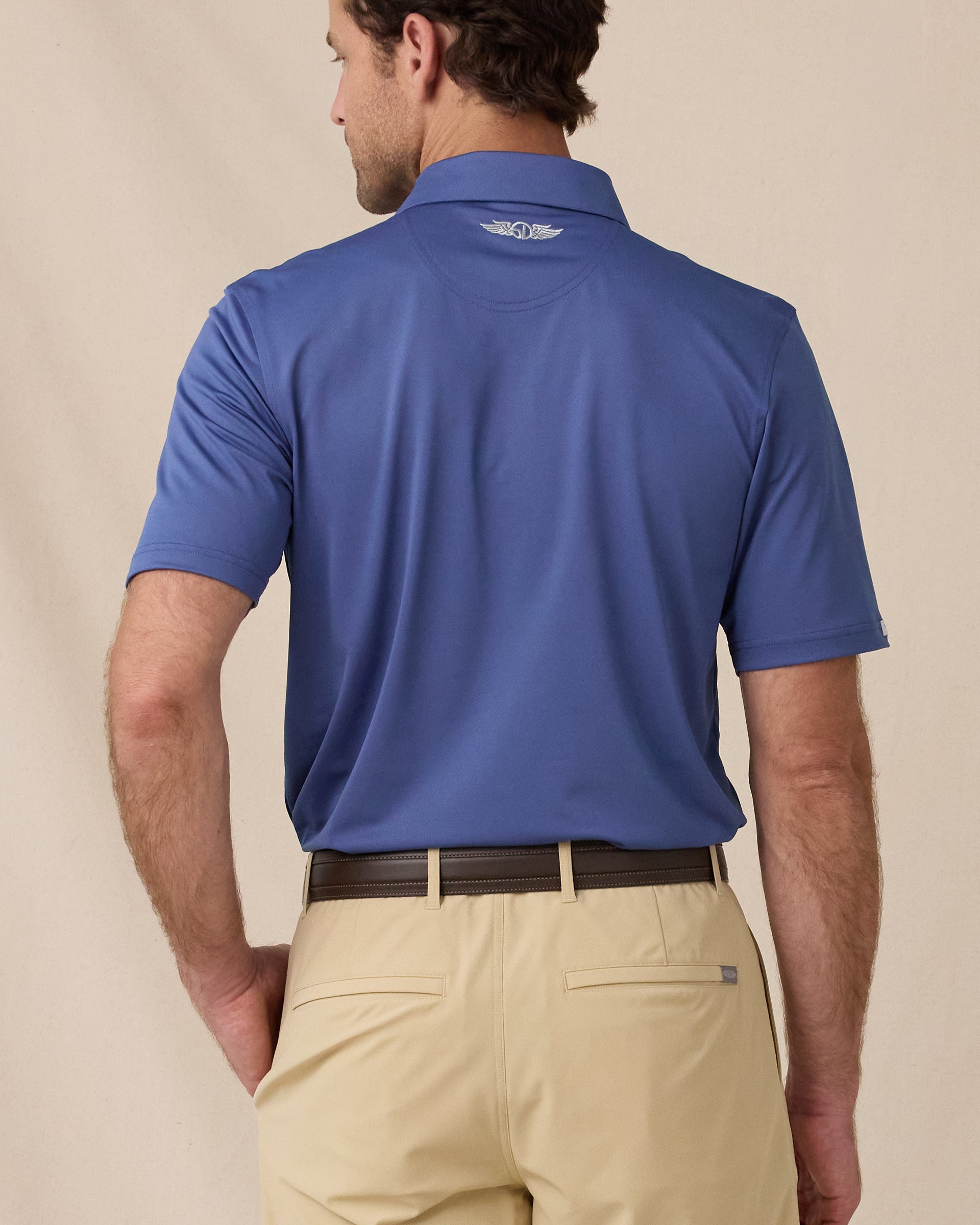 A model shown from the back wearing a blue short-sleeve polo shirt and khaki shorts, photographed against a plain beige studio background.