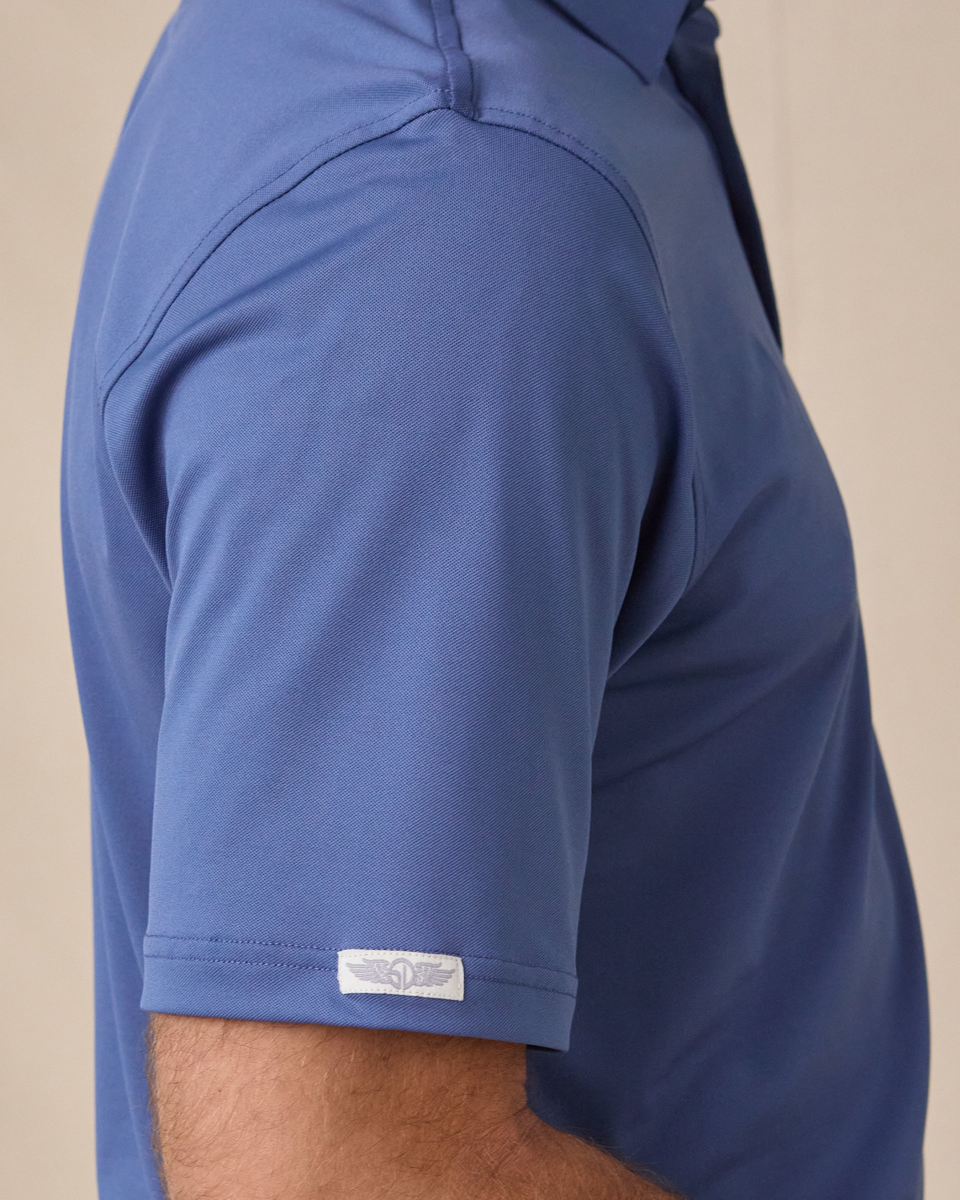 Close-up side view of a person wearing a short-sleeve polo shirt in a medium blue color, showing the sleeve cuff with a small white Straight Down wings tag.