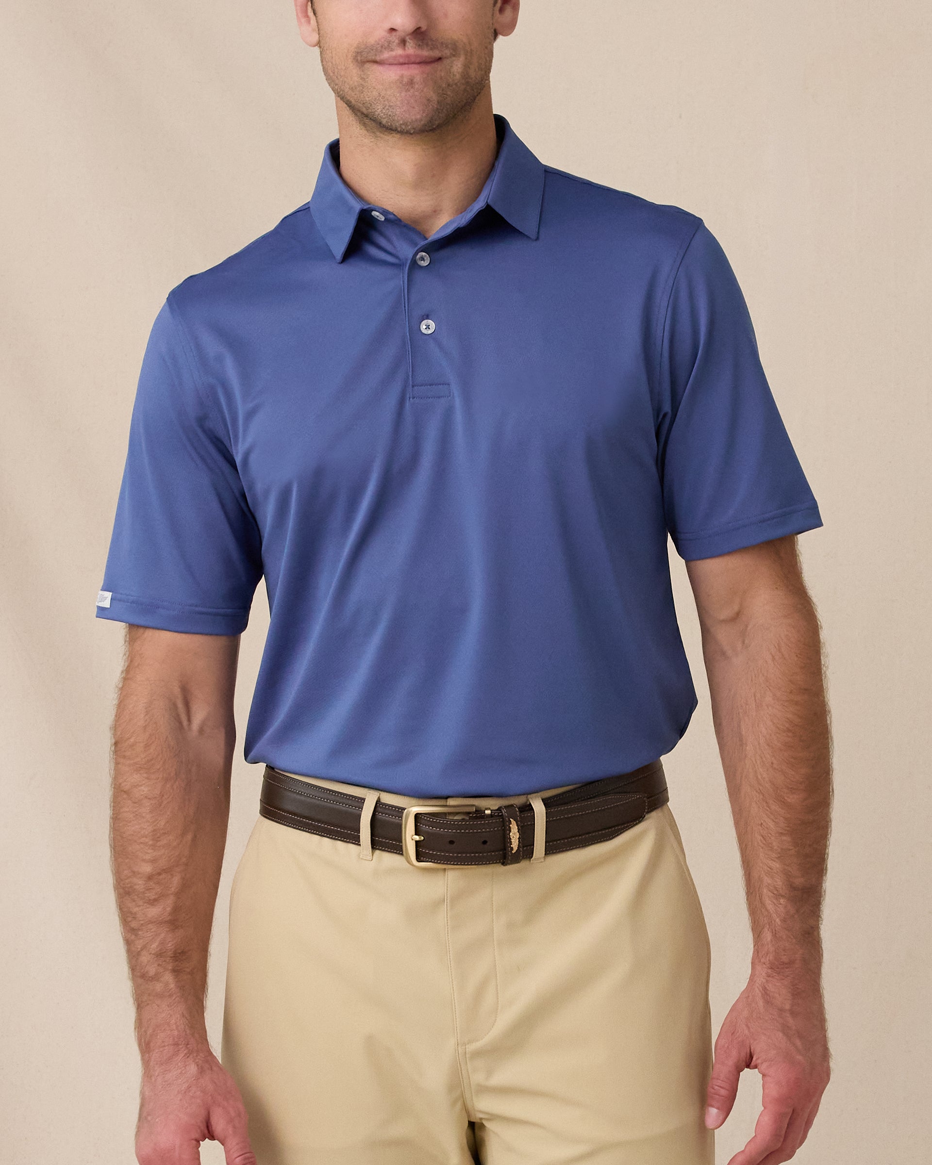 A model wearing a short-sleeve blue polo shirt with beige shorts and a dark brown belt, photographed against a plain beige studio background.