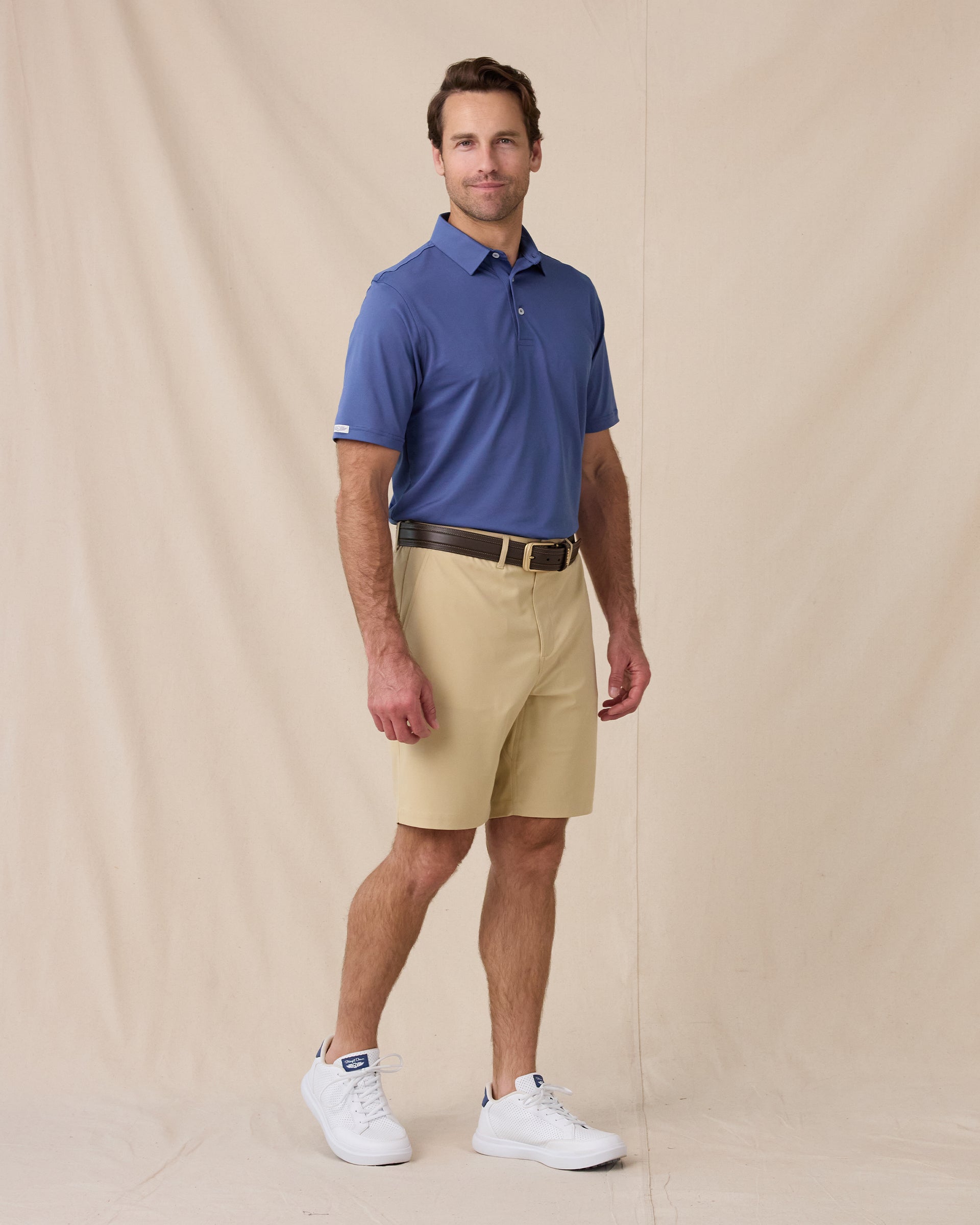 A man modeling a blue short-sleeve polo shirt and beige shorts against a beige fabric backdrop.