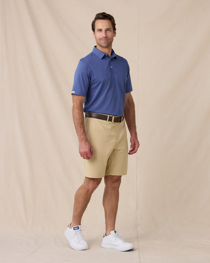 A man modeling a blue short-sleeve polo shirt and beige shorts against a beige fabric backdrop.