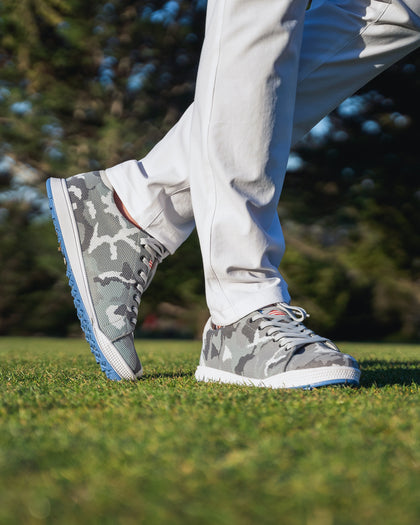 Men's Field V2 Shoe in Camo Carbon, on-course on-model view featuring the gray camo knit upper with white midsole and blue outsole, styled with white pants on a grass surface