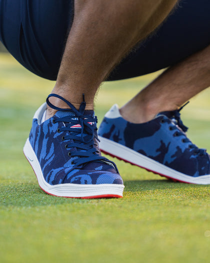 Men's Field V2 Shoe in Camo Indigo, on-course lifestyle view featuring a golfer crouching on the green wearing navy and blue camo print shoes with a white midsole and red outsole