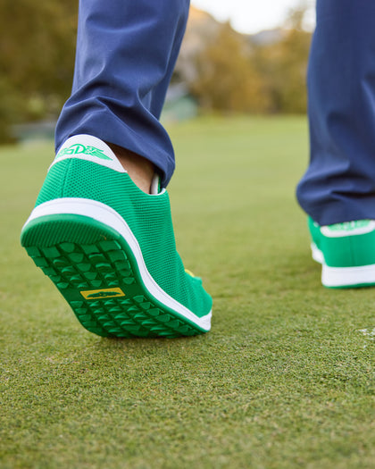 Men's Field V2 Shoe in Green, on-course lifestyle view featuring a close-up of a golfer walking on the green showing the green rubber outsole with multi-directional traction lugs and a yellow branded logo badge