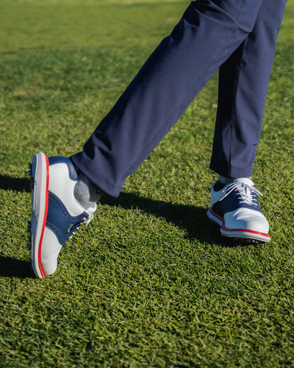 Men's Victory Classic Shoe in Red White Blue, on-course on-model view featuring the white leather upper with navy saddle overlay and red midsole welt, styled with navy pants on a grass surface