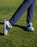 Men's Victory Classic Shoe in Red White Blue, on-course on-model view featuring the white leather upper with navy saddle overlay and red midsole welt, styled with navy pants on a grass surface