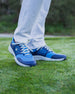 Men's Moonlight Shoe in Scuba, on-course on-model view featuring the light blue knit upper with navy overlays and white laces, styled with white pants on a grass surface