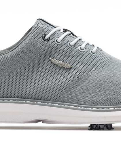 A close-up side view of a light gray knit golf shoe with white laces, a white sole, and a small silver wing logo on the side.