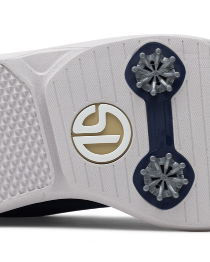 Men's Challenger Shoe in Indigo, close-up outsole view featuring a white rubber outsole with navy spike cleats and a gold and white branded logo badge detail