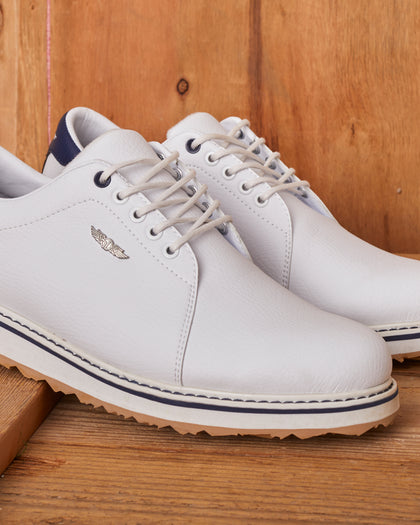 Straight Down Bourbon Hybrid Golf Shoe in White, angled lifestyle pair view on wood surface featuring white pebbled leather upper, navy heel counter, silver Straight Down wing logo badge, white laces, and navy stripe midsole accent with gum traction outsole