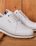 Straight Down Bourbon Hybrid Golf Shoe in White, angled lifestyle pair view on wood surface featuring white pebbled leather upper, navy heel counter, silver Straight Down wing logo badge, white laces, and navy stripe midsole accent with gum traction outsole