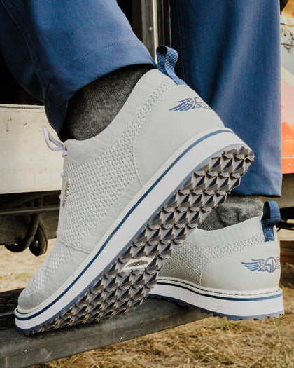 Straight Down Harbour Knit Hybrid Golf Shoe in Silver, lifestyle on-foot view featuring light gray knit upper, navy Straight Down wing logo on heel, and navy stripe midsole accent, styled with navy pants