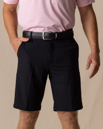 Men's Drake Short in Black, front model view featuring a flat-front silhouette styled with a black belt and pink polo