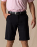 Men's Drake Short in Black, front model view featuring a flat-front silhouette styled with a black belt and pink polo