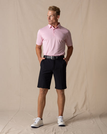 Men's Drake Short in Black, full-length front model view styled with a black belt, pink polo, and white golf shoes against a neutral canvas backdrop