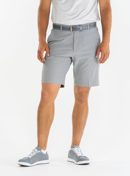 Men's Drake Short in Carbon, front model view featuring a flat-front silhouette in a cool medium gray colorway, styled with a gray belt, white polo, and gray golf shoes