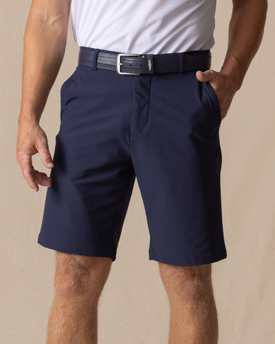 Men's Drake Short in Indigo, front model view featuring a flat-front silhouette in a rich navy blue colorway, styled with a navy belt and white polo