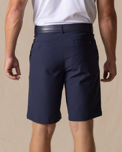 Men's Drake Short in Indigo, back model view featuring button-welt back pockets with tonal logo detail in a rich navy blue colorway, styled with a navy belt and white polo
