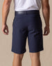 Men's Drake Short in Indigo, back model view featuring button-welt back pockets with tonal logo detail in a rich navy blue colorway, styled with a navy belt and white polo