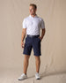 Men's Drake Short in Indigo, full-length front model view in a rich navy blue colorway, styled with a navy belt, white polo, and white golf shoes against a neutral canvas backdrop