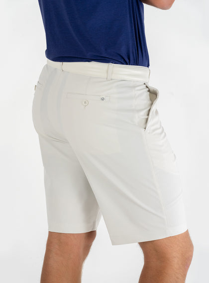 Men's Drake Short in Stone, back model view featuring a button-welt back pocket with tonal logo detail in a warm off-white colorway, styled with a white belt and navy polo